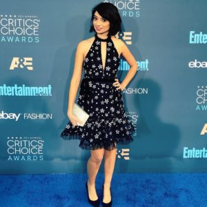 Kate Micucci in a black dress with her arm on her hip holding a clutch